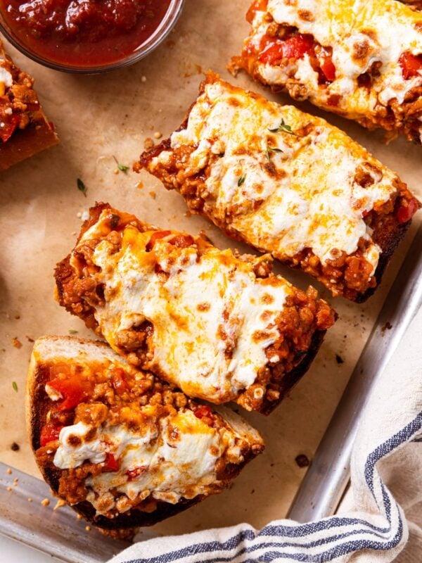 Baked slices of sausage French bread pizza sitting on brown piece of parchment on baking sheet with melted cheese over the top showing bits of tomato sauce and sausage.
