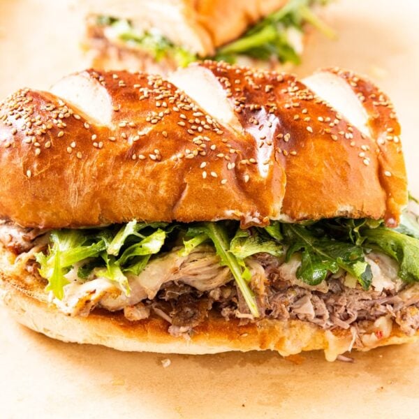 Half of a roasted pork sandwich topped with greens and cheese on French bread sitting on a piece of brown-colored parchment paper on white surface.