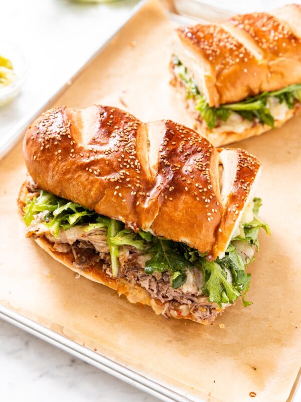 Parchment-lined baking sheet with two halves of Italian roasted pork sandwich with greens and cheese on top of meat in between slices of French bread with greens and pepperoncini in background.
