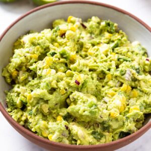 White marble surface with brown bowl filled with sweet corn guacamole with chunky texture.