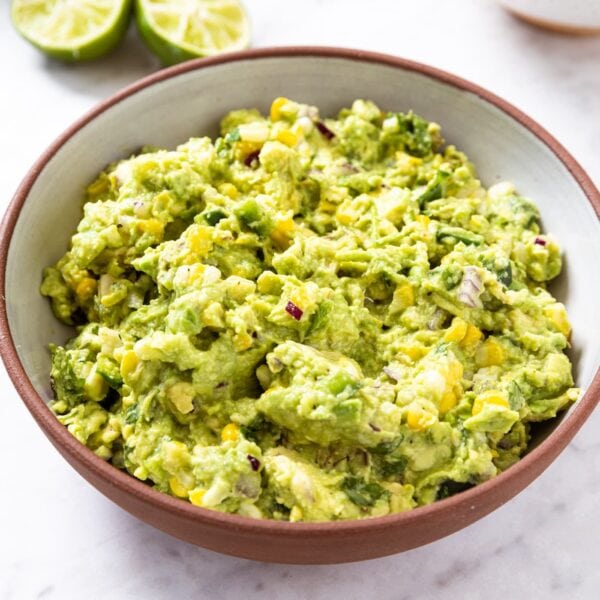 White marble surface with brown bowl filled with sweet corn guacamole with sliced lime and lemon in background.
