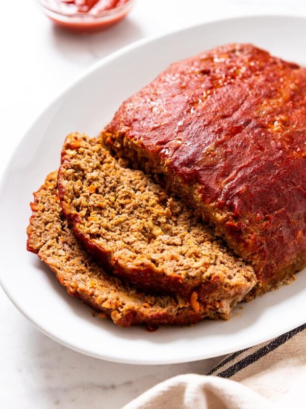 White serving platter with meatloaf topped with ketchup mixture with two slices resting against side of platter with small glass bowl filled with ketchup all on marble surface.