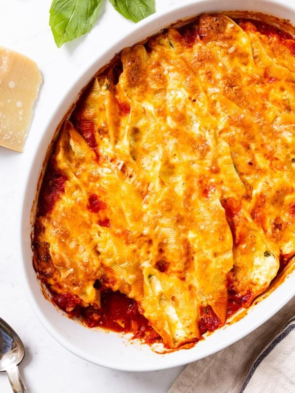 White marble surface with oval white baking dish filled with stuffed shells with cheese shredded on top sitting on a bed of tomato sauce.