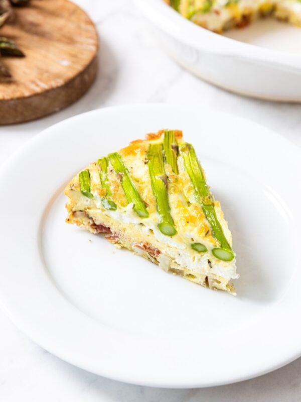 White plate with slice of roasted potato frittata with pieces of asparagus baked on the top of the slice with wood board in background.