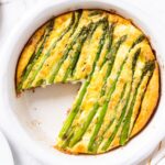 White pie dish filled with baked asparagus and roasted potato frittata with stalks of asparagus all over the top baked into the egg filling on white marble surface.