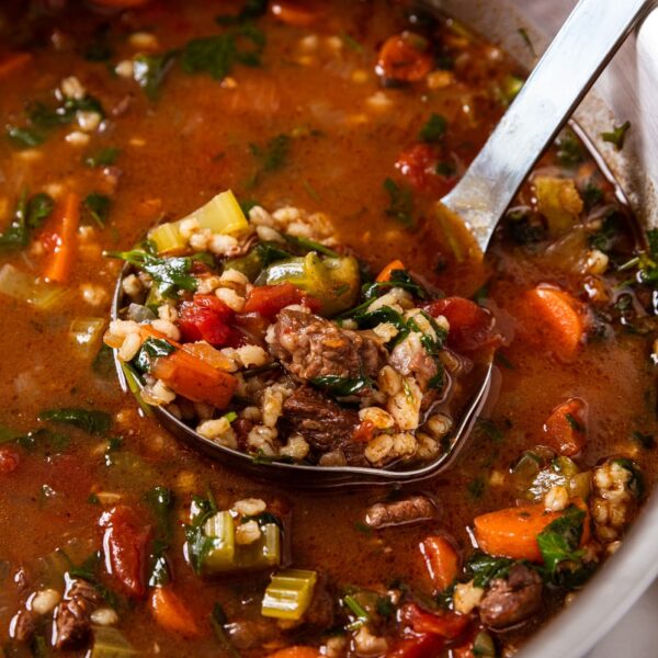 Large metal stockpot sitting on white marble surface with metal ladle resting on the bottom of the pot with beef and barley resting in the ladle.