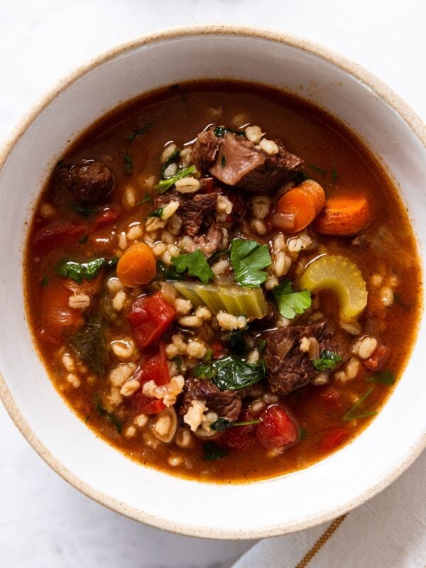 White stoneware bowl filled with brown-colored beef and barley soup with pieces of beef, carrot, and celery all on white marble surface.