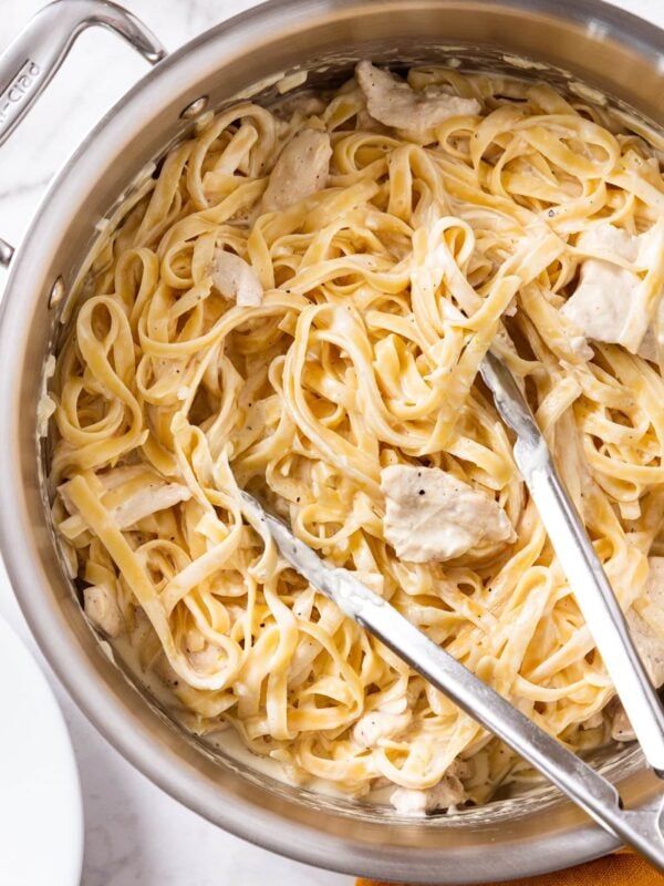 Large metal stockpot filled with fettuccine alfredo with chicken mixed into creamy sauce with tongs resting on side of pot.