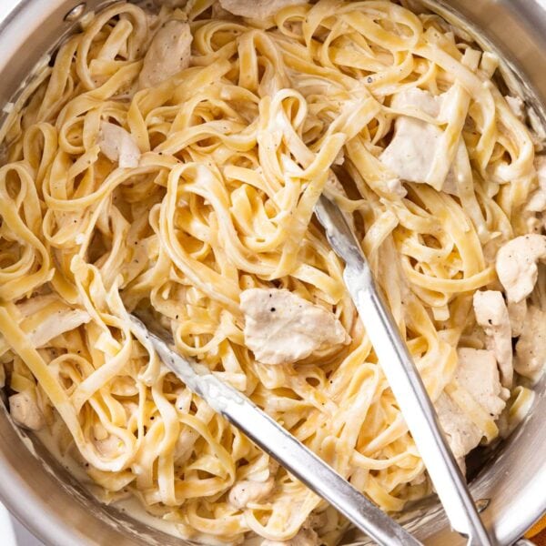 Large metal stockpot filled with fettuccine alfredo with pieces of chicken resting on top along with tongs all on white marble surface.