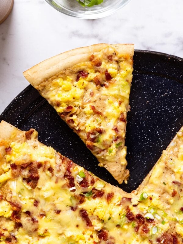 Black pizza pan with bacon breakfast pizza topped with eggs resting on marble surface with one slice missing.