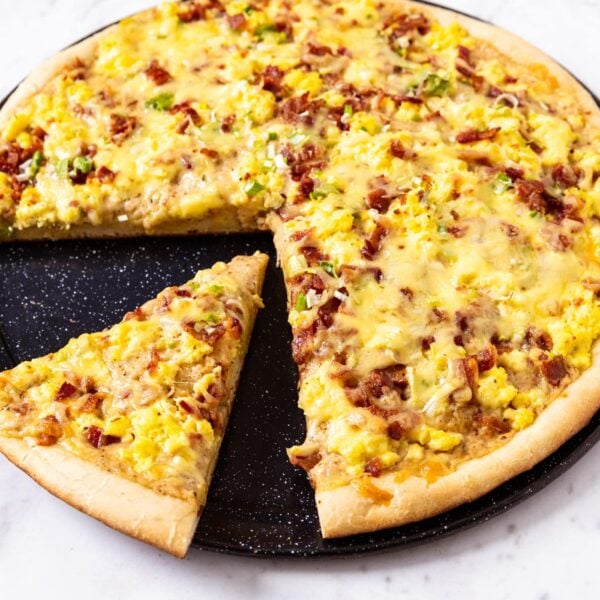 Black pizza pan holding baked bacon breakfast pizza with eggs, cheese, and chopped scallion all over the top with one slice of pizza removed from the rest.