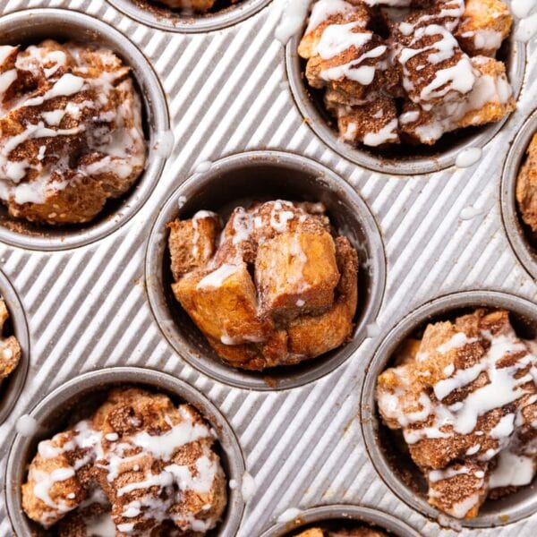 Metal gray muffin pan filled with baked muffins with thin glaze drizzled over the top with one muffin turned on its side to see the interior texture.