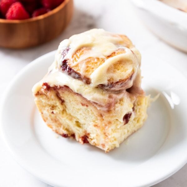 White plate with one raspberry cinnamon roll sitting on it frosted with cream cheese frosting with bowl of raspberries and rest of cinnamon rolls in background.