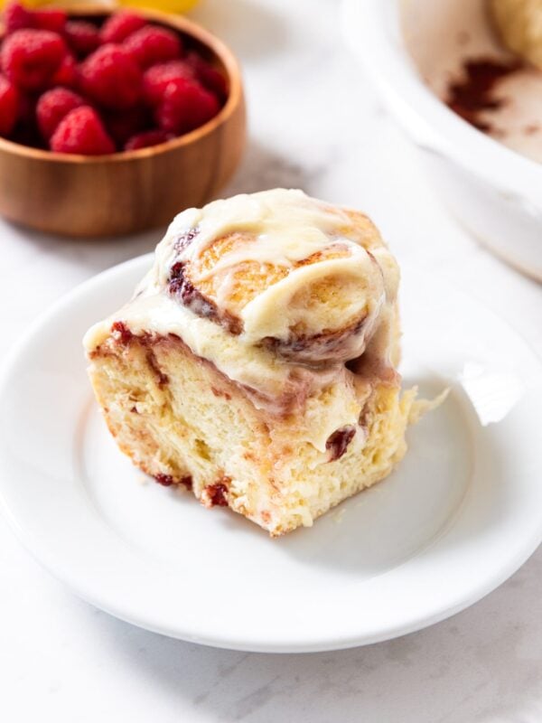 White plate with single raspberry cinnamon roll sitting on it with bowl of raspberries in background along with lemons and rest of baking dish of cinnamon rolls.