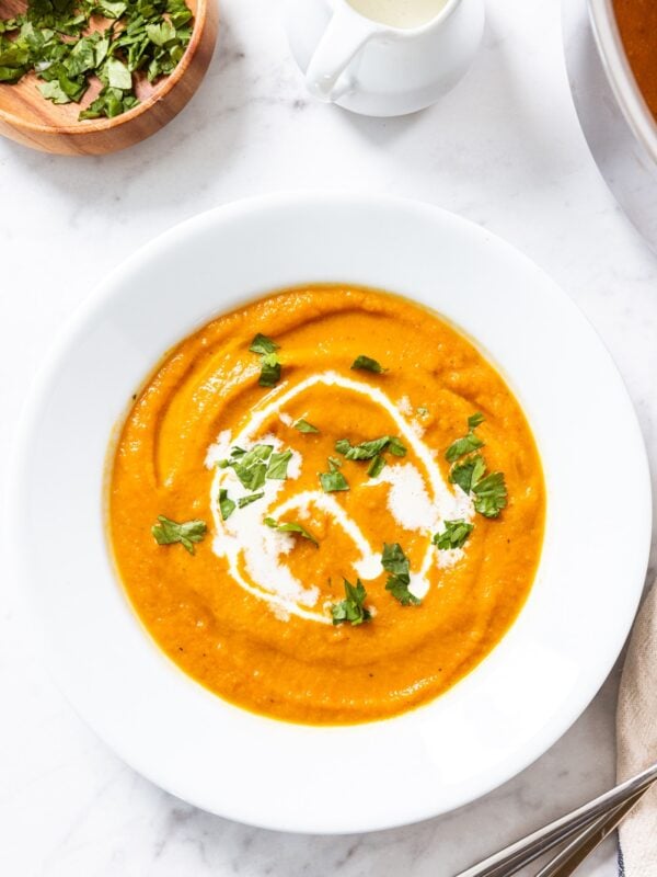White bowl sitting on marble surface filled with blended roasted carrot soup topped with drizzle of heavy cream and chopped cilantro with rest of soup in stockpot to the side.