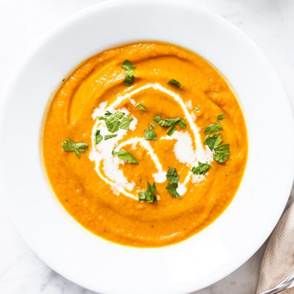 White plate bowl filled with blended orange-colored roasted carrot soup with swirl of heavy cream and chopped cilantro on top sitting on white marble surface.