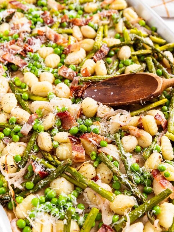 Sheet pan filled with gnocchi, asparagus, peas, and sliced shallots with wooden spoon resting on side of sheet pan.