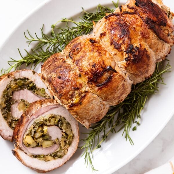 White marble surface filled with white platter with a cooked pork roulade sitting on a bed of herbs with two slices showing the interior swirl filled with kale, onion, and fennel mixture.