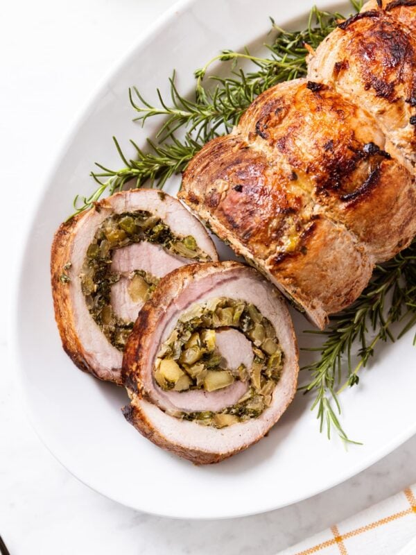 White platter filled with cooked pork roulade with two slices cut off the end showing the interior swirl filled with onion, fennel, and kale filling.