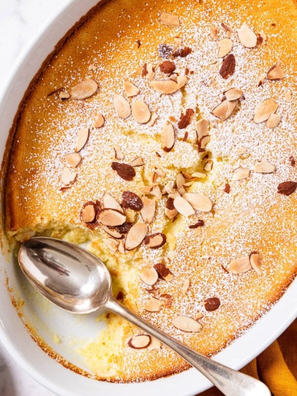 Oval baking dish filled with baked lemon pudding cake topped with a dusting of powdered sugar and toasted almonds with a spoon resting on side of bowl after removing a serving.