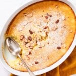 Oval baking dish sitting on marble surface filled with baked lemon pudding cake dusted with powdered sugar and topped with toasted almonds with spoon resting on side of platter after removing a serving.