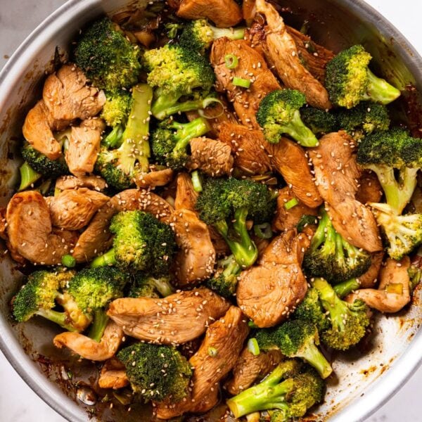 White marble surface with metal skillet filled with cooked sesame chicken and broccoli with soy sauce in bottom pan with small bowl filled with sesame seeds to the side.