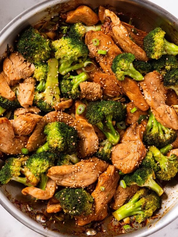 White marble surface with metal skillet filled with cooked pieces of chicken and broccoli florets tossed in soy sauce with sesame seeds all over the top.