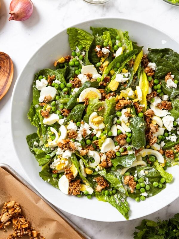 White serving platter filled with spring pea salad with creamy dressing all over the top along with crunchy nut topping all on white marble surface.