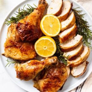 White circular platter filled with sliced pieces of citrus roast chicken with two halves of citrus laying open along with herbs all on white marble surface.