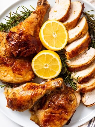 White circular platter filled with sliced pieces of citrus roast chicken with two halves of citrus laying open along with herbs all on white marble surface.