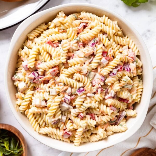 Marble surface with large white bowl filled with creamy pasta salad with red bell pepper and hamburger buns to the side.