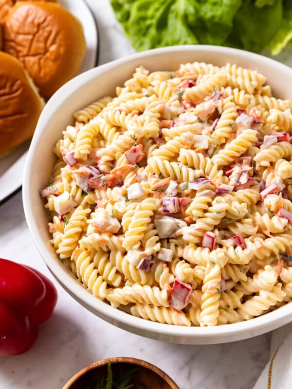 Large white bowl filled with creamy pasta salad with lettuce and sandwich buns in background all on white marble surface.