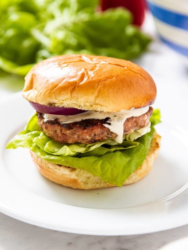 White plate sitting on marble surface with grilled pork burger with creamy mayonnaise sauce dripping down the side of the meat with lettuce under the meat and more lettuce in background.