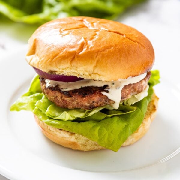 White plate sitting on marble surface with grilled pork burger sitting with a bit of creamy mayonnaise dressing about to drip down the side with more lettuce and red onion in the background.