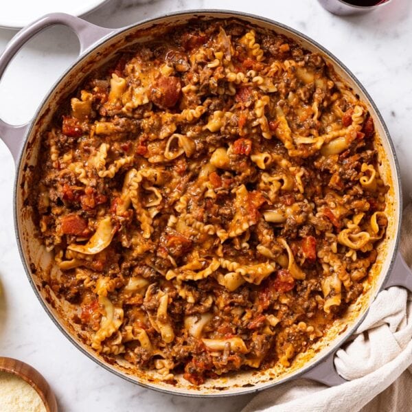 White marble surface filled with large pan filled with ragù sauce with pasta showing along with olive oil, Parmesan cheese, and cups of red wine.