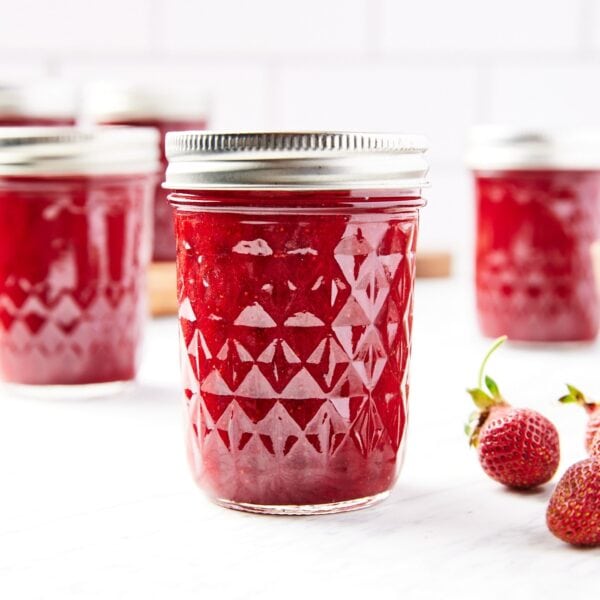 Glass jam jars filled with strawberry butter sitting on marble surface with tile background with lids and rings on top.