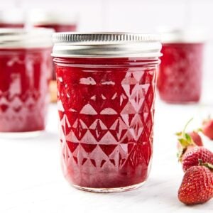 Glass jam jars filled with red colored strawberry butter sitting on marble surface with lids and rings on top.