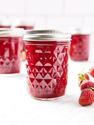 Glass jam jars filled with red colored strawberry butter sitting on marble surface with lids and rings on top.