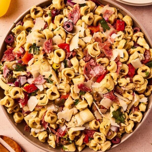 Large brown bowl filled with Italian tortellini pasta salad with red onion, bell pepper, artichoke hearts, parsley, olives, and more all on taupe surface.