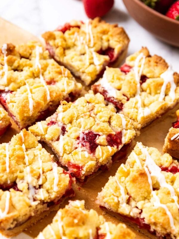 Strawberry crumb bars topped with white glaze sitting on piece of brown parchment paper with more strawberries in background.