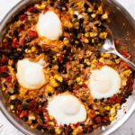 White marble surface with metal skillet filled with tomato, sweet corn, and black bean mixture with cooked soft eggs on top with spoon resting on the side of skillet.
