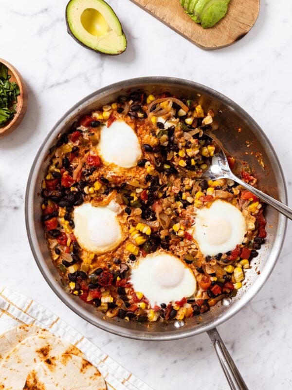 White marble surface with metal skillet filled with tomato, bean, and corn mixture with eggs cooked right on top with tortillas and avocado to put on top to the side.