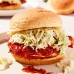 Barbecue chicken sandwich sitting on tan colored plate with layer of coleslaw on top of layer of barbecue with pieces spilling onto the plate.