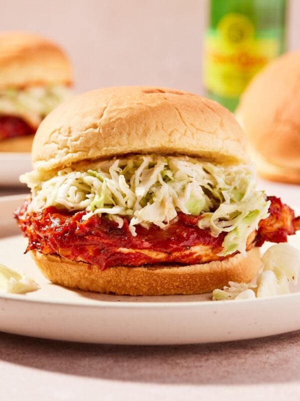 Barbecue chicken sandwich with layer of coleslaw sitting on tan colored plate with bottle of sparkling water in background and more sandwich buns.