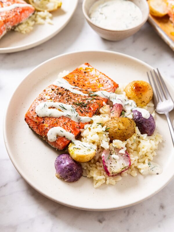 White marble surface with tan colored plate with salmon filet and roasted radishes over a bed of cooked rice and topped with ranch dressing with more ranch dressing in the background.