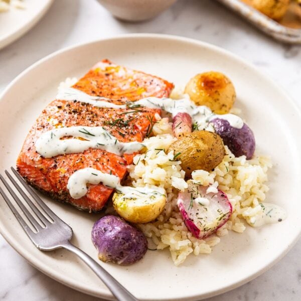 White marble surface with bed of white rice topped with filet of cooked salmon and sliced radishes and drizzled with homemade ranch dressing with baking sheet in background.