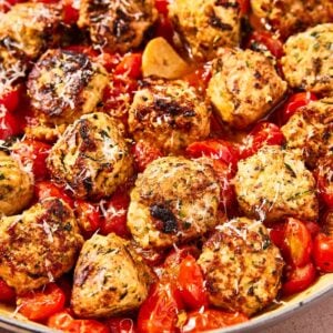 Large skillet filled with chicken zucchini meatballs nestled in tomato sauce with burst grape tomatoes and pieces of garlic after cooking.