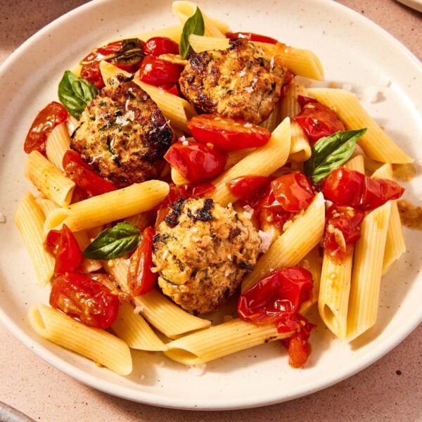 White plate with penne topped with cooked tomatoes and chicken zucchini meatballs with pieces of basil and a sprinkle of salt on top.