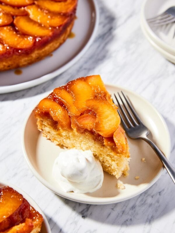 White marble surface with taupe colored plate with slice of upside down peach cake with dollop of whipped cream to the side.