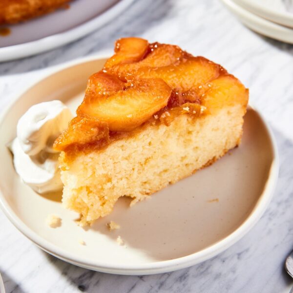 White marble surface with taupe-colored plate with slice of upside down peach cake with dollop of whipped cream to the side.